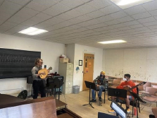 Two guitar students seated on the right side of the classroom, a guitar instructor stands at the front of the room holding a lute