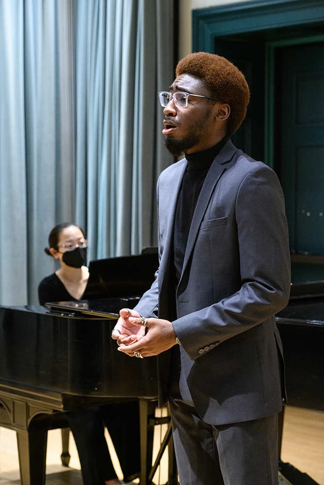 A male vocalists performing in the foreground with a pianist in the background