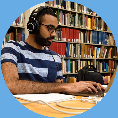 A student sits at a table with headphones on and plugged into a laptop. A bookshelf is seen behind the student